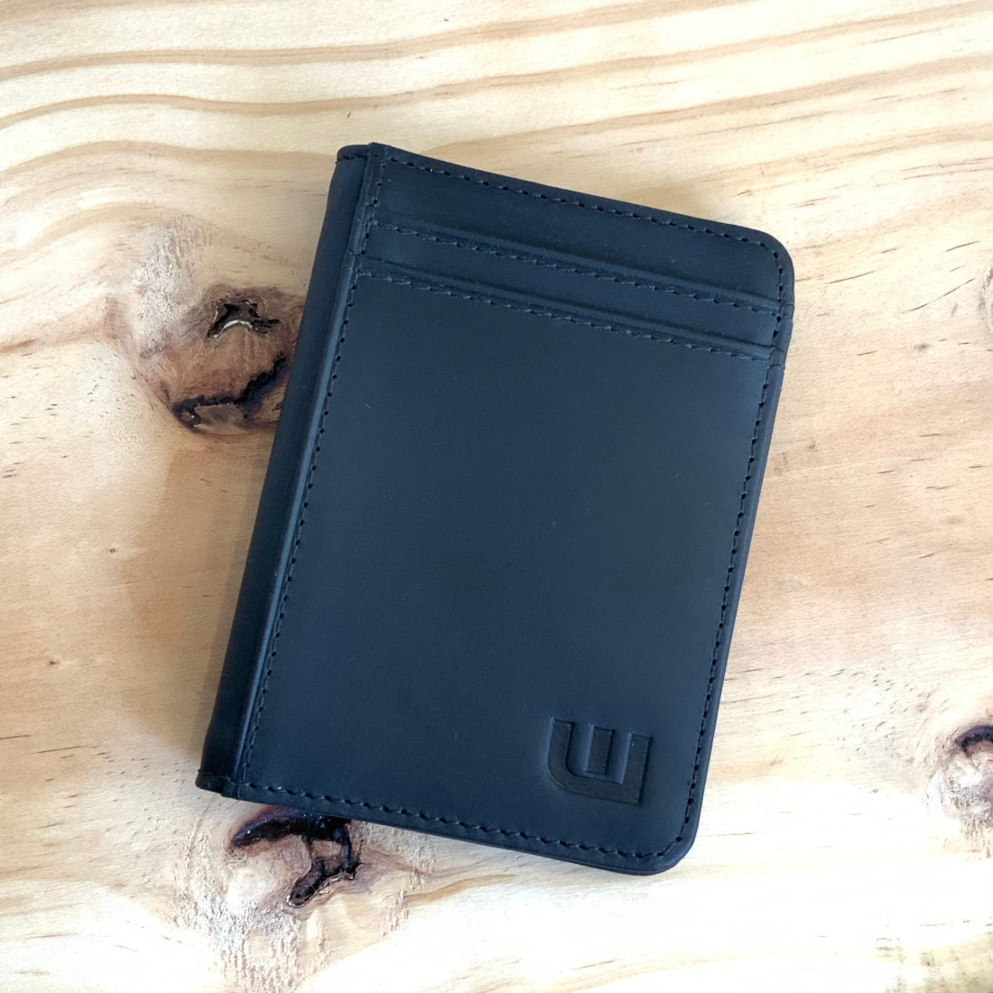 A black leather card holder wallet with a debossed logo, placed on a light wood grain surface.