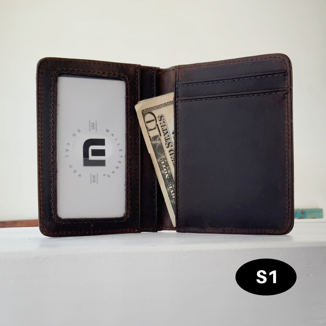 A dark brown leather bifold wallet shown open, with an ID window on the left, card slots on the right, and a dollar bill partially tucked inside. A black oval label reading 'S1' appears in the lower right corner.