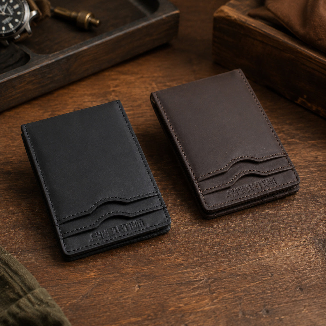 Two slim cardholder wallets, one black and one dark brown, displayed side-by-side on a rustic wooden table with a watch and leather goods in the background.