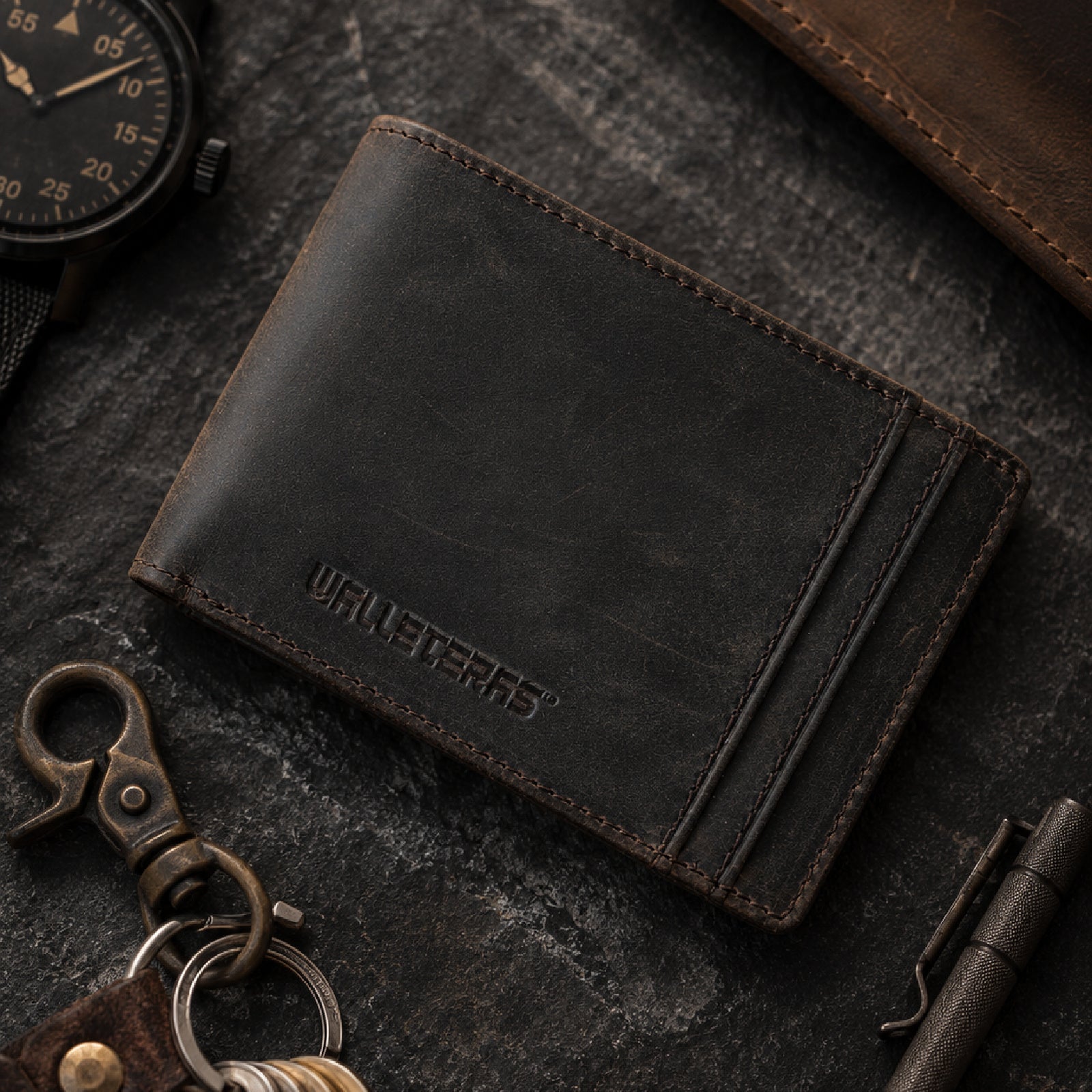 A dark brown distressed leather bifold wallet styled on a dark textured surface with a watch, keychain, and pen.